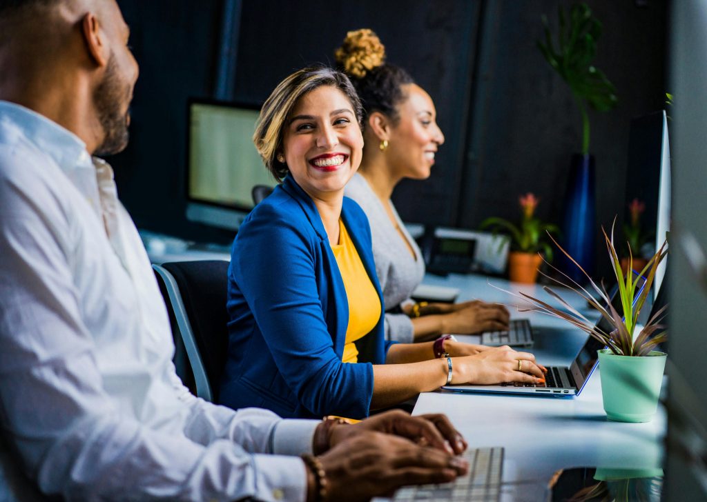 pexels-photo-2422293-2422293 Three diverse professionals working and smiling at office desks, fostering teamwork and collaboration.